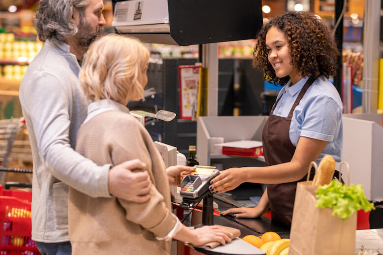 Happy female cashier looking at mature couple paying for what they bought by credit card in supermarket