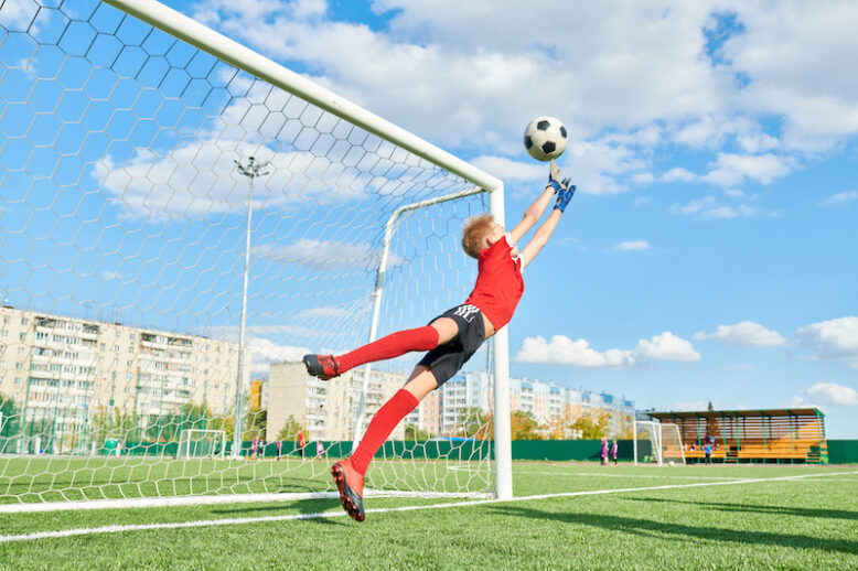 Full length portrait of teenage boy jumping forward and catching ball while defending gate in football match