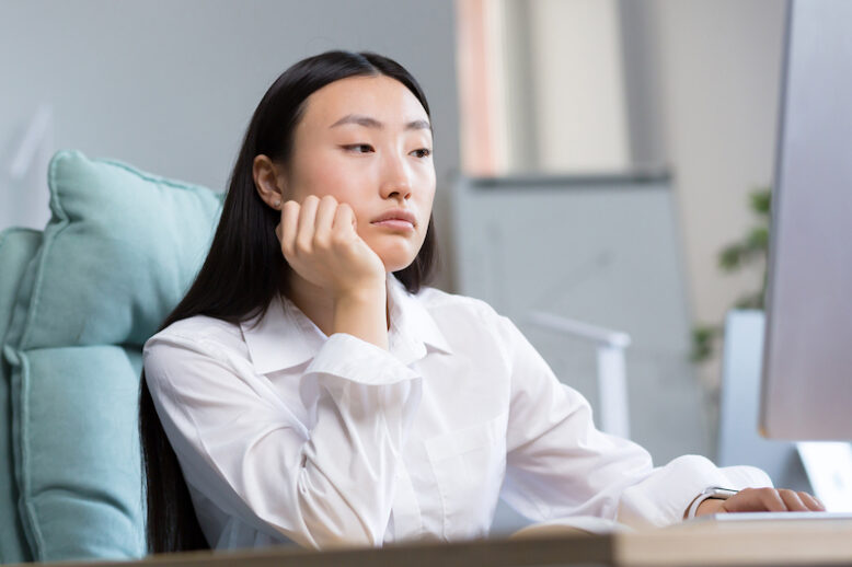 Boring and monotonous work. A young beautiful Asian business woman, an office worker sits at a desk at a computer, is sad, bored, performs monotonous work. He holds his head with one hand.