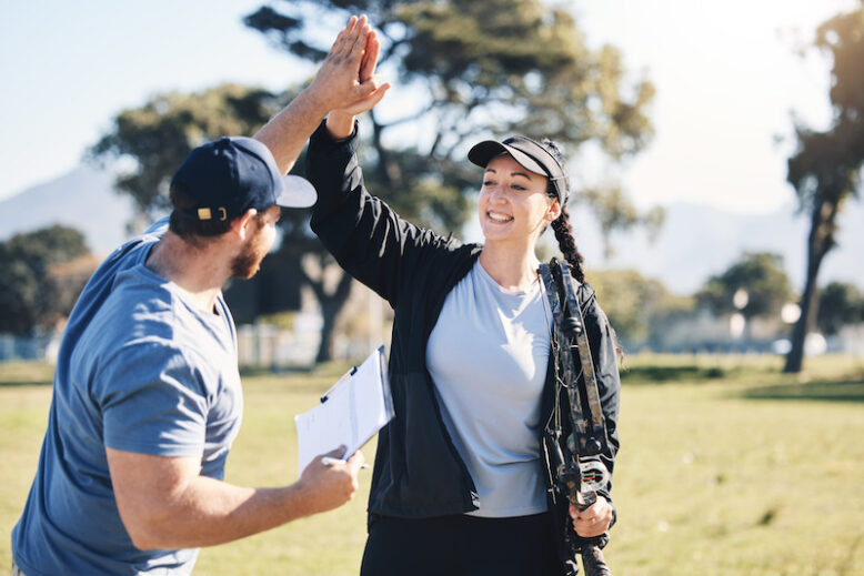 Woman, archery and personal trainer high five in sports for partnership, success or celebration on .