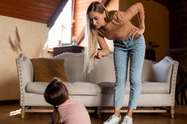 Family Conflicts And Punishment Concept. Strict Angry Mother Scolding Her Upset Scared Little Daughter For Her Behaviour While She Sitting On The Floor At Home. Naughty Kid, Children Abuse