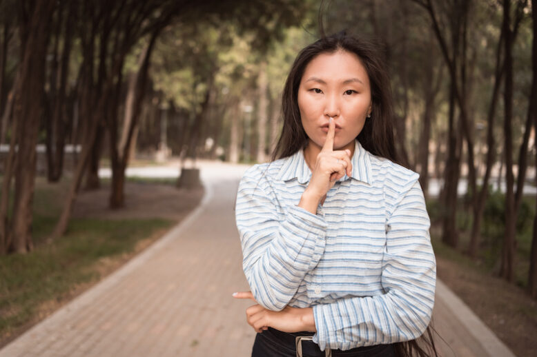 Young beautiful Asian woman holding index finger on her mouth lips standing in forest. be hush silence